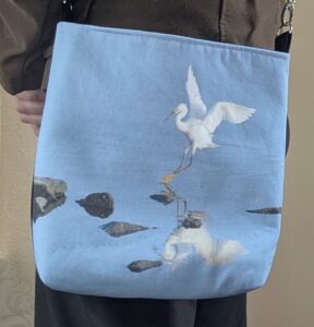 A durable weight cotton crossbody bag featuring a white Snowy Egret bird landing with upturned wings in shallow blue water with rocks. The egret is alsoreflected in the blue water. The bag has an adjustable strap with silver-toned hardware.